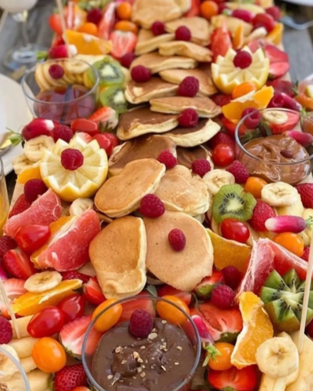 Grande table garnie de mini-pancakes, fruits frais découpés et coupelles de pâte à tartiner
