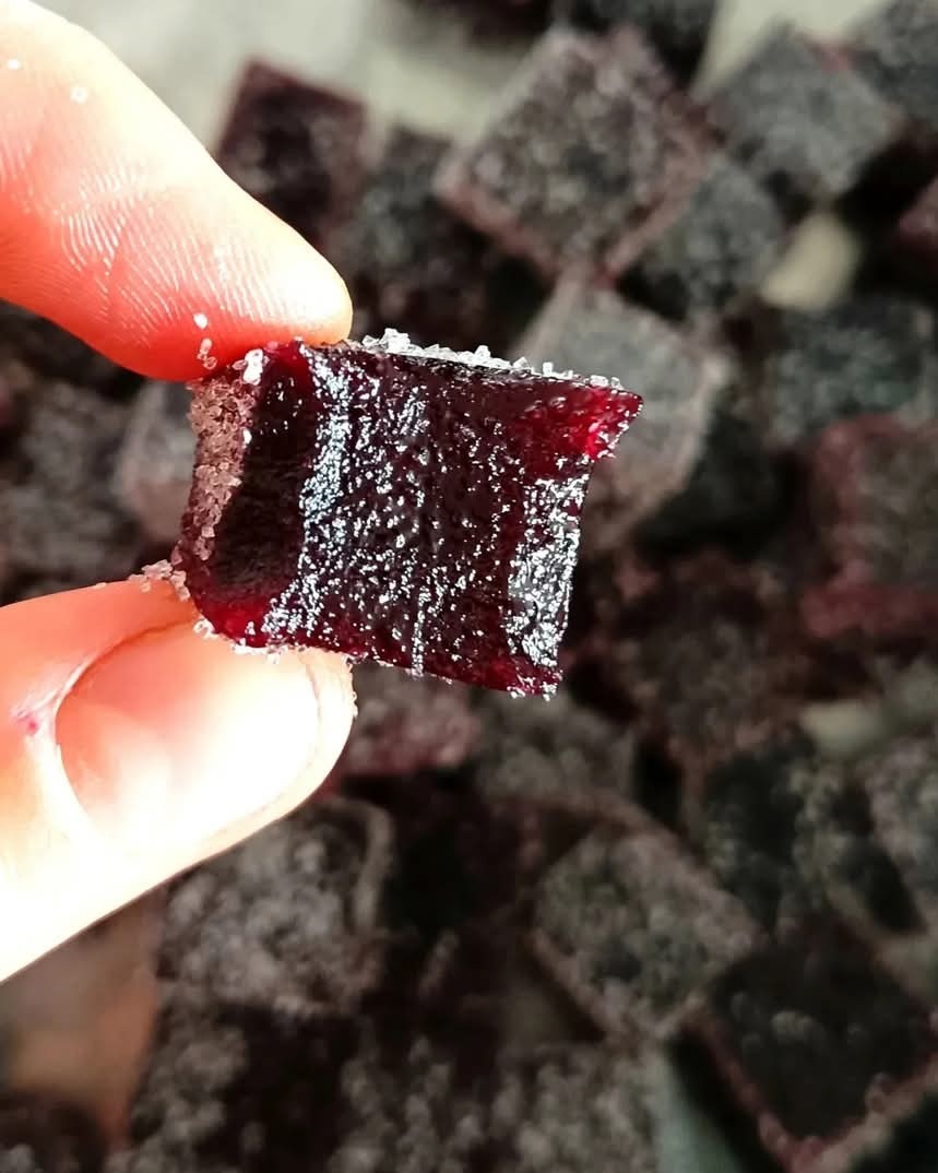 Main tenant une pâte de fruits rouge enrobée de sucre, devant un plateau de pâtes de fruits