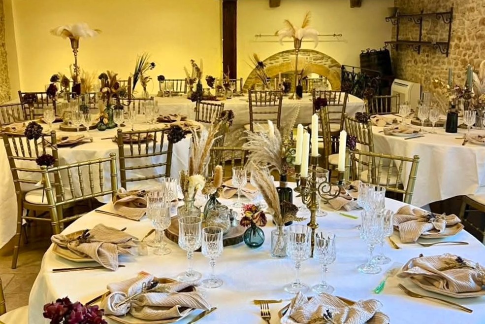 Salle de réception en pierre, tables rondes dressées de nappes blanches, chaises dorées, bougies et bouquets de pampa