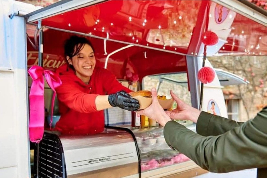 Depuis le comptoir de son camion, une cuisinière de L'Épicurienne tend un burger dans une barquette à un client