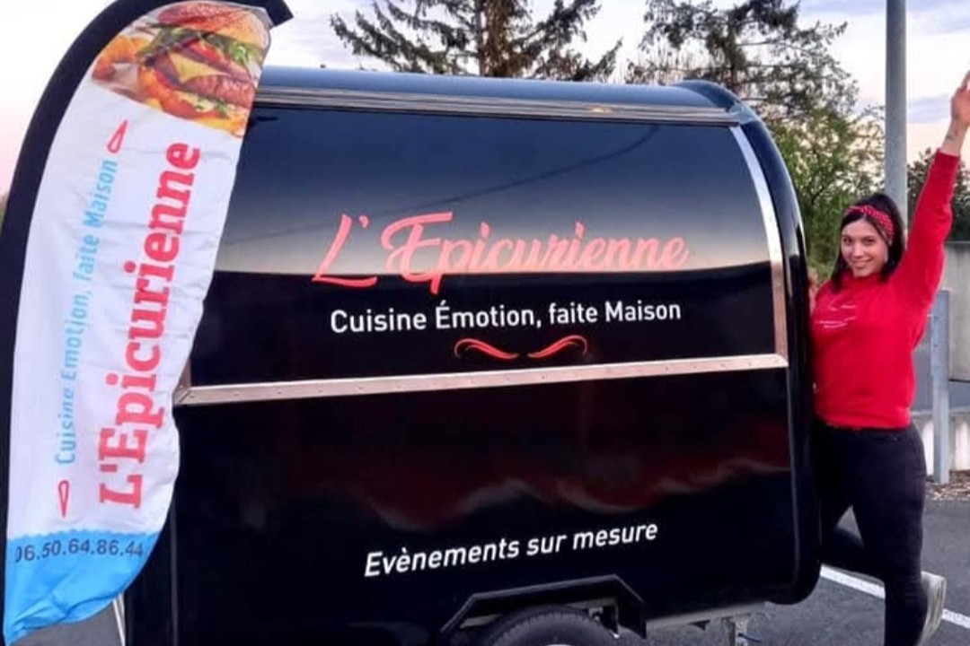 Océane, bras levé, devant la Mini-Roulotte noire floquée « L'Épicurienne — Cuisine Émotion, faite Maison — Évènements sur mesure »